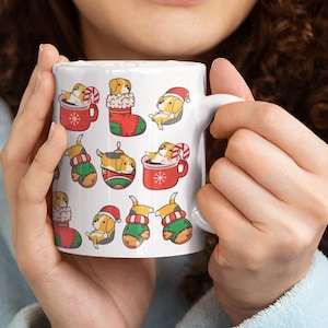 May include: White ceramic mug with a Christmas-themed design featuring a pattern of dogs wearing Santa hats and Christmas sweaters. The mug is held by a person in front of a Christmas tree.