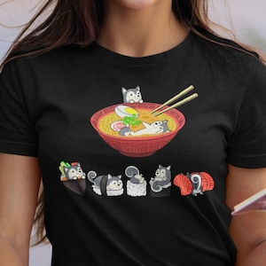 May include: A black t-shirt featuring a graphic of cartoon dogs enjoying ramen and sushi. The design shows a bowl of ramen with a dog inside, and a line of sushi with dogs on top.