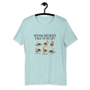 Pu&ograve; includere: Una maglietta blu chiaro con una grafica di un pastore tedesco e il testo "German Shepherd's Daily To-Do List". La grafica mostra il cane in diverse pose, tra cui dormire, fare rumore, stirarsi, distruggere qualcosa, osservare gli umani e dormire ancora.