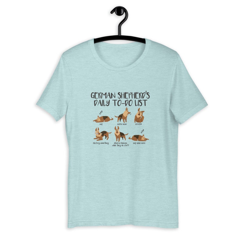 Pu&ograve; includere: Una maglietta blu chiaro con una grafica di un pastore tedesco e il testo "German Shepherd's Daily To-Do List". La grafica mostra il cane in diverse pose, tra cui dormire, fare rumore, stirarsi, distruggere qualcosa, osservare gli umani e dormire ancora.