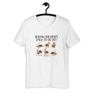 Pu&ograve; includere: Maglietta bianca con un'illustrazione di un pastore tedesco e il testo "German Shepherd's Daily To-Do List". L'illustrazione mostra il cane che dorme, fa rumore, si gratta, distrugge qualcosa, distrae gli umani mentre fanno cose e dorme ancora.