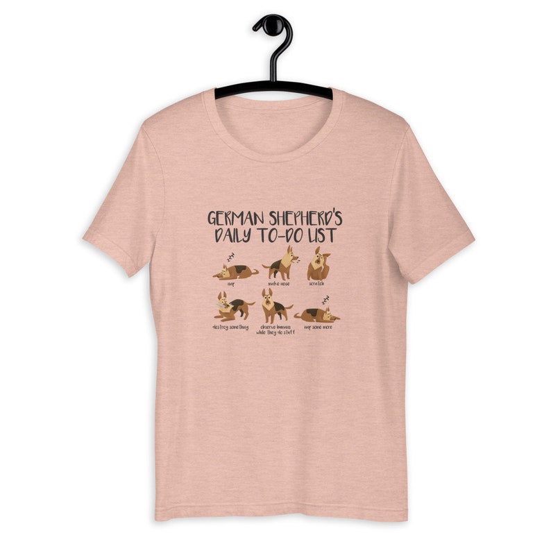 Pu&ograve; includere: Una t-shirt rosa melange con una grafica di un pastore tedesco in varie pose. Il testo "GERMAN SHEPHERD'S DAILY TO-DO LIST" &egrave; stampato sopra la grafica. La grafica include il testo "nap", "make a mess", "yawn", "destroy something", "observe humans while they do their stuff" e "nap some more".