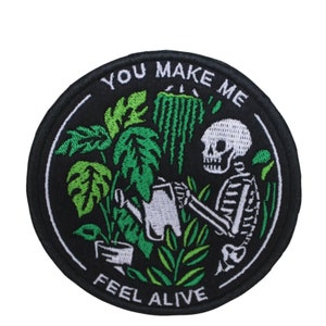 May include: A black and white embroidered patch with a skeleton watering plants. The patch is round and has the text "You Make Me Feel Alive" around the edge.