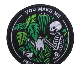 May include: A black and white embroidered patch with a skeleton watering plants. The patch is round and has the text "You Make Me Feel Alive" around the edge.