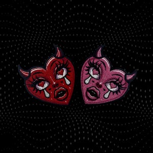 Peut inclure: Deux écussons brodés en forme de cœur. L'un est rouge, l'autre rose. Chacun présente un visage avec de longs cils, une larme et des cornes de diable. Les cœurs ont une bordure noire et sont posés sur un fond noir à pois.