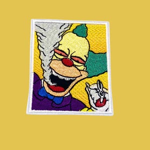 May include: Embroidered patch featuring a cartoon clown with a green hat, purple bow tie, and a red nose. The clown is smiling and appears to be smoking a cigarette. The patch has a white border.