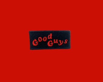 May include: A black rectangular patch with the embroidered text "Good Guys" in red script. The patch is set against a solid red background, creating a striking contrast.