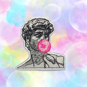 Peut inclure: Un patch brodé représentant une sculpture célèbre soufflant une bulle rose. Le patch est sur un fond coloré.