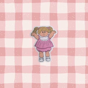 May include: A sticker illustration of a doll with blonde hair in pigtails, wearing a pink dress with a white collar and pink bloomers. The doll has a round face, tan skin, and is depicted with arms raised. The sticker has a white border outline.