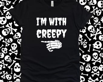 May include: Black t-shirt featuring the white text "I'M WITH CREEPY" and a skeleton hand pointing left. The shirt has a crew neck design. The background is a pattern of white skulls and crossbones.