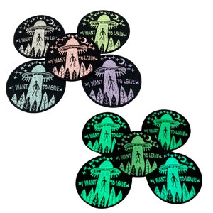 May include: Eight black and white embroidered patches with a green, pink, or blue glow-in-the-dark design. Each patch features a UFO with a person standing in the doorway and the text "I WANT TO LEAVE".
