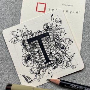 Op de afbeelding: Een zwart-wit Zentangle kunstwerk met de letter "T" omringd door bloemen en abstracte ontwerpen. Het kunstwerk staat op een vierkant, gebroken wit papier, met een potlood en pen op de voorgrond. Het papier heeft de tekst "an original zentangle".