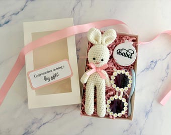 May include: A gift box containing a cream-colored crocheted rabbit with a pink ribbon, a round white mirror with the name Isabel printed in black, and sunglasses with white daisy-shaped frames. The box lid features the text Congratulations on being a big sister.
