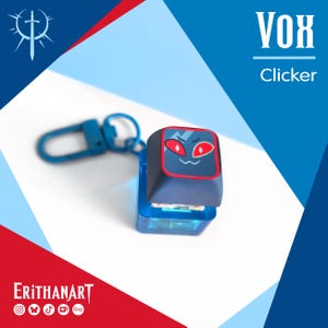 Peut inclure: Un porte-clés interrupteur de clavier mécanique bleu avec une touche gris foncé ornée d'un graphique rouge représentant un visage aux yeux rouges et une petite bouche. Le texte VOX Clicker est visible en haut à droite, ERITHANART en bas à gauche.