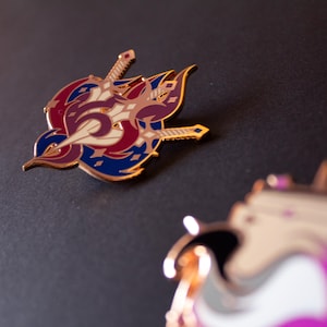 May include: A detailed enamel pin featuring a stylized design with swords and a crest in shades of red, blue, cream, and gold. The pin has a metallic gold border and is set against a dark background. The design is intricate and eye-catching.