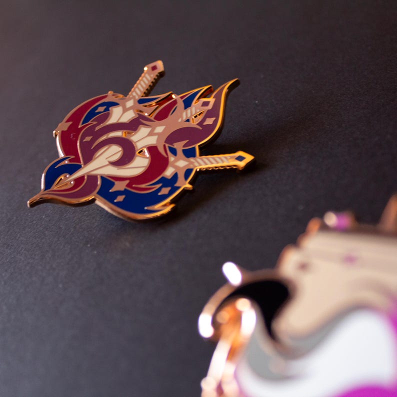 May include: A detailed enamel pin featuring a stylized design with swords and a crest in shades of red, blue, cream, and gold. The pin has a metallic gold border and is set against a dark background. The design is intricate and eye-catching.
