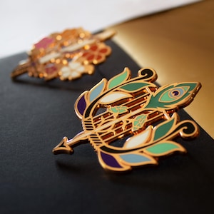 May include: Two decorative gold-colored pins. The front pin is shaped like a bow and arrow with green, blue, purple, and white enamel details. The second pin, out of focus, has a floral design with pink, white, and orange accents.