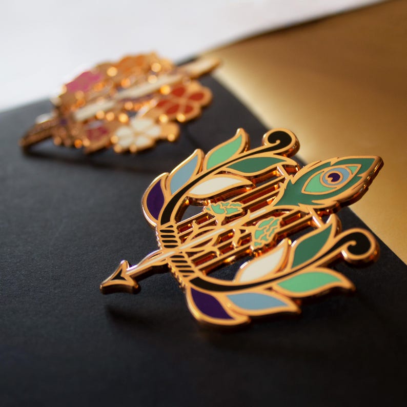 May include: Two decorative gold-colored pins. The front pin is shaped like a bow and arrow with green, blue, purple, and white enamel details. The second pin, out of focus, has a floral design with pink, white, and orange accents.