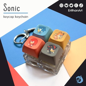 Può includere: Un portachiavi con quattro tasti colorati con personaggi di Sonic the Hedgehog. I tasti sono blu, arancioni, gialli e grigi, ognuno con un design diverso. Il portachiavi include un anello di metallo argentato e il testo "Sonic keycap keychain".
