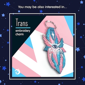 May include: An embroidered charm with a design featuring pink, blue, and white colors. The charm has a key ring and the word "Trans" is displayed above the words "embroidery charm". The charm has an eye in the center.