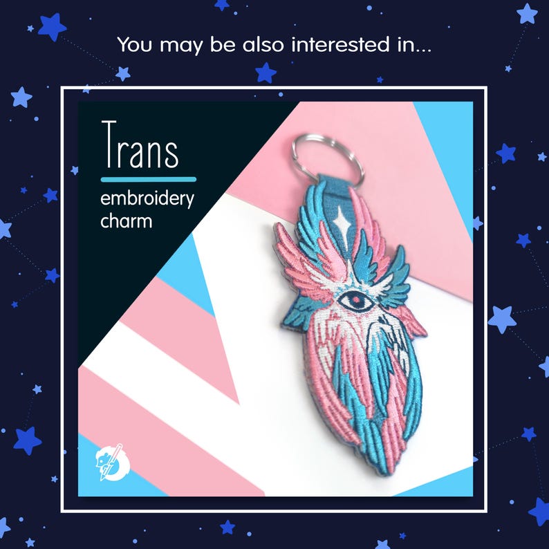 May include: An embroidered charm with a design featuring pink, blue, and white colors. The charm has a key ring and the word "Trans" is displayed above the words "embroidery charm". The charm has an eye in the center.