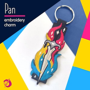 Peut inclure: Porte-clés brodé avec un motif d'épée, avec des flammes jaunes, bleues, roses et blanches. Le porte-clés a un dos noir et un porte-clés argenté. Le texte sur l'image indique "Pan embroidery charm."