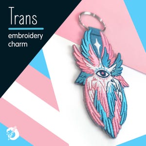 Peut inclure: Un porte-clés brodé avec un motif aux couleurs rose, bleu et blanc. Le charm présente un motif stylisé d'œil et de plumes. Les mots "Trans" et "broderie charme" sont imprimés sur le fond.