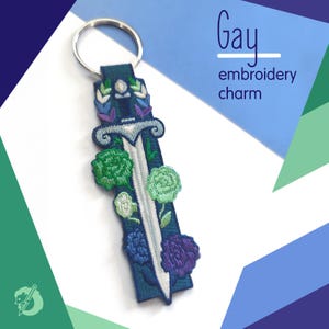Può includere: Un portachiavi ricamato rettangolare blu scuro con un design a spada argentata decorato con motivi floreali verdi, verde chiaro, blu e viola. Il testo "Gay embroidery charm" è stampato nell'angolo in alto a destra dell'immagine.