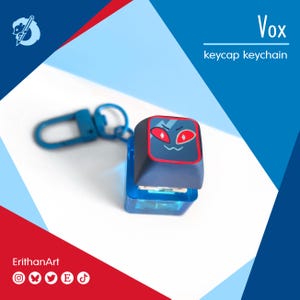 May include: A blue keycap keychain with a cartoon face design. The keycap is dark blue with a red and black face. The keychain has a blue clasp and the text "Vox keycap keychain".
