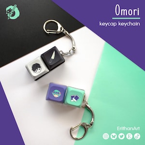 May include: Two keycap keychains featuring Omori branding. One keychain has a white keycap with a character face and a black keycap with a knife icon. The other has a purple keycap with a flower-crowned character and a mint green keycap with two photos.