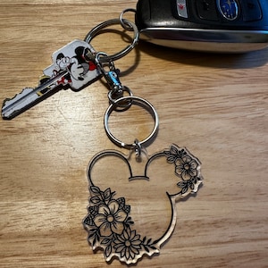 K&ouml;nnte beinhalten: Ein durchsichtiger Acryl-Schl&uuml;sselanh&auml;nger mit einer schwarzen Umrandung von Mickey Mouse-Ohren mit Blumendesigns. Der Schl&uuml;sselanh&auml;nger ist an einem silbernen Schl&uuml;sselring befestigt.
