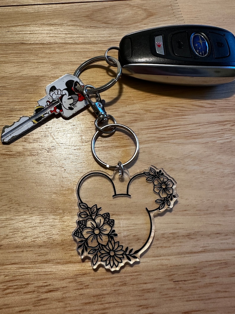 K&ouml;nnte beinhalten: Ein durchsichtiger Acryl-Schl&uuml;sselanh&auml;nger mit einer schwarzen Umrandung von Mickey Mouse-Ohren mit Blumendesigns. Der Schl&uuml;sselanh&auml;nger ist an einem silbernen Schl&uuml;sselring befestigt.