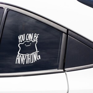 May include: A white vinyl decal applied to a car window featuring a stylized, amorphous blob character with a simple line face. The text "YOU CAN BE ANYTHING" is written in a bold, white, hand-lettered font above and below the character illustration.