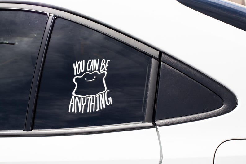 May include: A white vinyl decal applied to a car window featuring a stylized, amorphous blob character with a simple line face. The text "YOU CAN BE ANYTHING" is written in a bold, white, hand-lettered font above and below the character illustration.