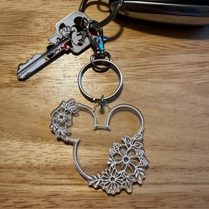 K&ouml;nnte beinhalten: Ein wei&szlig;er Acryl-Schl&uuml;sselanh&auml;nger in Form von Mickey-Maus-Ohren mit wei&szlig;en Blumendetails. Der Schl&uuml;sselanh&auml;nger ist an einem silbernen Schl&uuml;sselring befestigt.