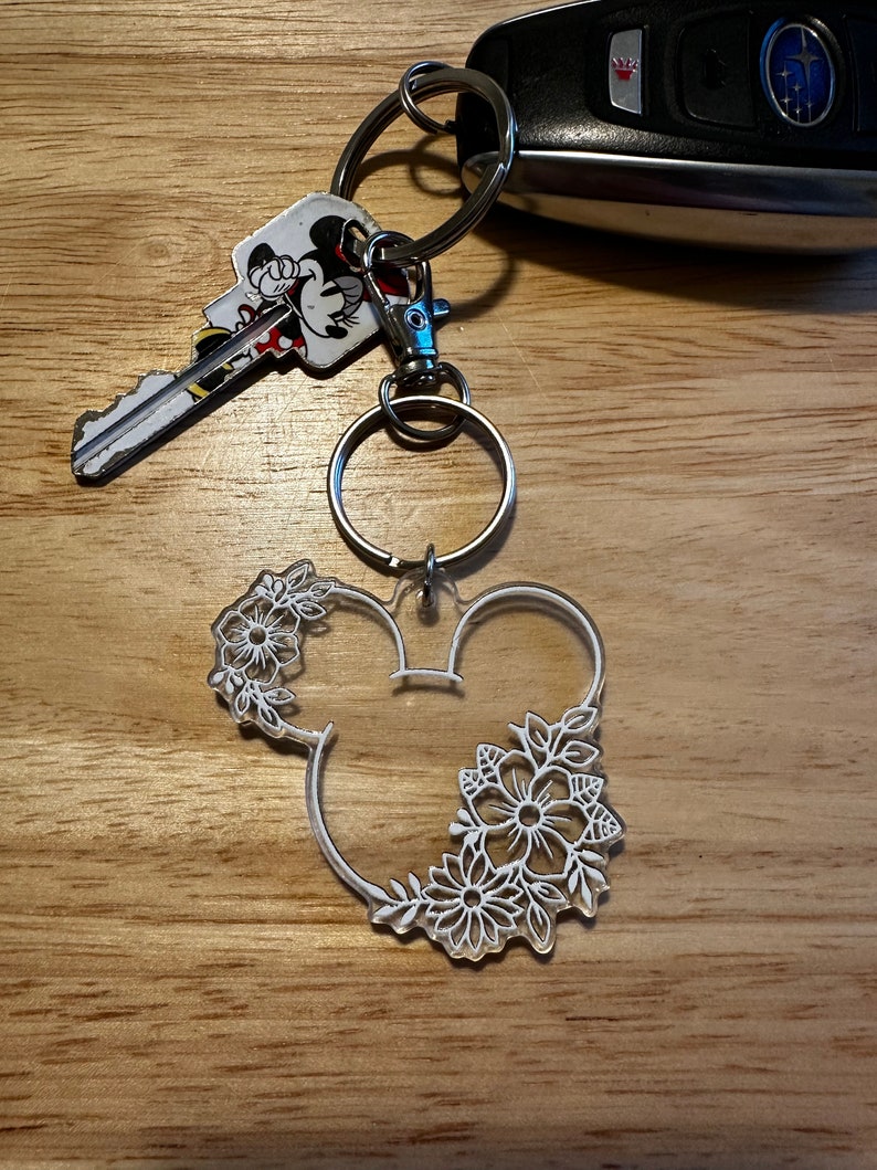 K&ouml;nnte beinhalten: Ein wei&szlig;er Acryl-Schl&uuml;sselanh&auml;nger in Form von Mickey-Maus-Ohren mit wei&szlig;en Blumendetails. Der Schl&uuml;sselanh&auml;nger ist an einem silbernen Schl&uuml;sselring befestigt.