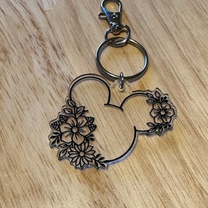 K&ouml;nnte beinhalten: Ein durchsichtiger Acryl-Schl&uuml;sselanh&auml;nger in Form von Mickey-Maus-Ohren mit schwarzen Blumenmustern. Der Schl&uuml;sselanh&auml;nger hat einen silbernen Metallring und einen silbernen Metallkarabiner.