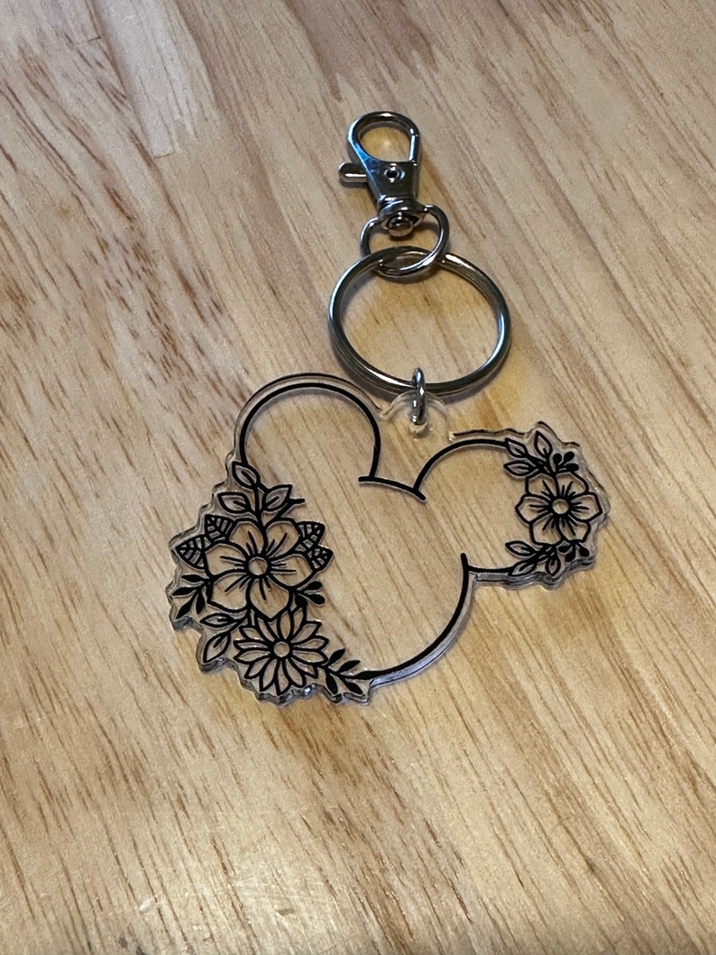 K&ouml;nnte beinhalten: Ein durchsichtiger Acryl-Schl&uuml;sselanh&auml;nger in Form von Mickey-Maus-Ohren mit schwarzen Blumenmustern. Der Schl&uuml;sselanh&auml;nger hat einen silbernen Metallring und einen silbernen Metallkarabiner.