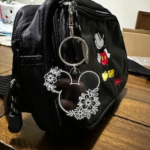 K&ouml;nnte beinhalten: Ein schwarzer Rucksack mit Mickey-Mouse-Motiv und einem wei&szlig;en, floralen Mickey-Mouse-Schl&uuml;sselanh&auml;nger, der an einem silbernen Ring befestigt ist.