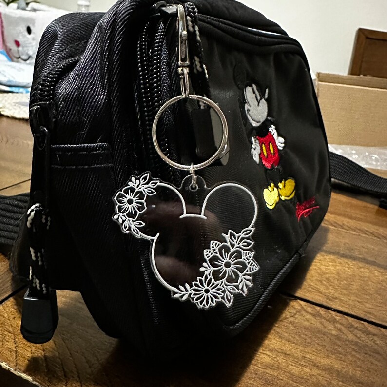 K&ouml;nnte beinhalten: Ein schwarzer Rucksack mit Mickey-Mouse-Motiv und einem wei&szlig;en, floralen Mickey-Mouse-Schl&uuml;sselanh&auml;nger, der an einem silbernen Ring befestigt ist.