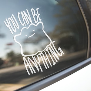 May include: A white vinyl decal applied to a car window featuring a line-art illustration of a blob-shaped character with a simple face and the text YOU CAN BE ANYTHING written in a clean, sans-serif font arching above and below the character.