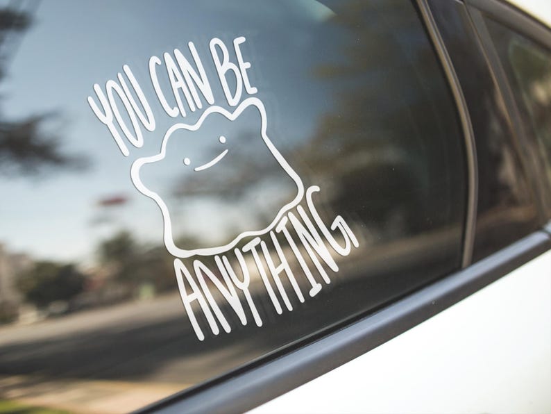 May include: A white vinyl decal applied to a car window featuring a line-art illustration of a blob-shaped character with a simple face and the text YOU CAN BE ANYTHING written in a clean, sans-serif font arching above and below the character.