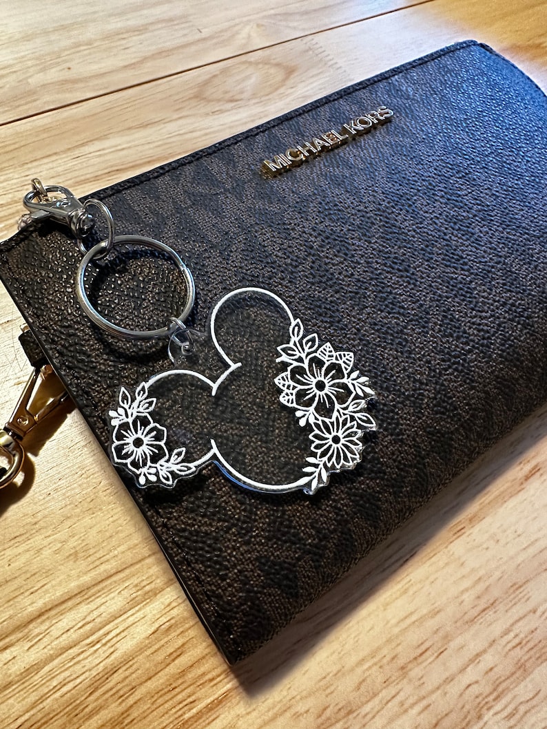 K&ouml;nnte beinhalten: Ein Schl&uuml;sselanh&auml;nger mit einem wei&szlig;en floralen Mickey-Mouse-Design, befestigt an einem silbernen Schl&uuml;sselring und einem goldenen Verschluss. Der Schl&uuml;sselanh&auml;nger h&auml;ngt an einer schwarzen Michael Kors-Handtasche.