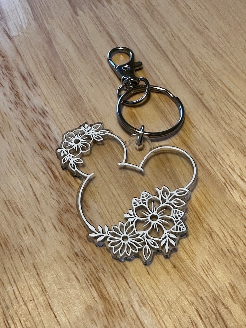 K&ouml;nnte beinhalten: Ein durchsichtiger Acryl-Schl&uuml;sselanh&auml;nger in Form von Mickey-Maus-Ohren mit einem wei&szlig;en Blumenmuster. Der Schl&uuml;sselanh&auml;nger hat einen silbernen Metallring und einen silbernen Metallverschluss.