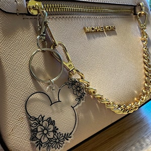 K&ouml;nnte beinhalten: Eine hellrosa Michael Kors Handtasche mit einer goldenen Kettenriemen und einem durchsichtigen Schl&uuml;sselanh&auml;nger mit einer schwarzen Umrandung von Mickey Mouse Kopf mit floralen Details.
