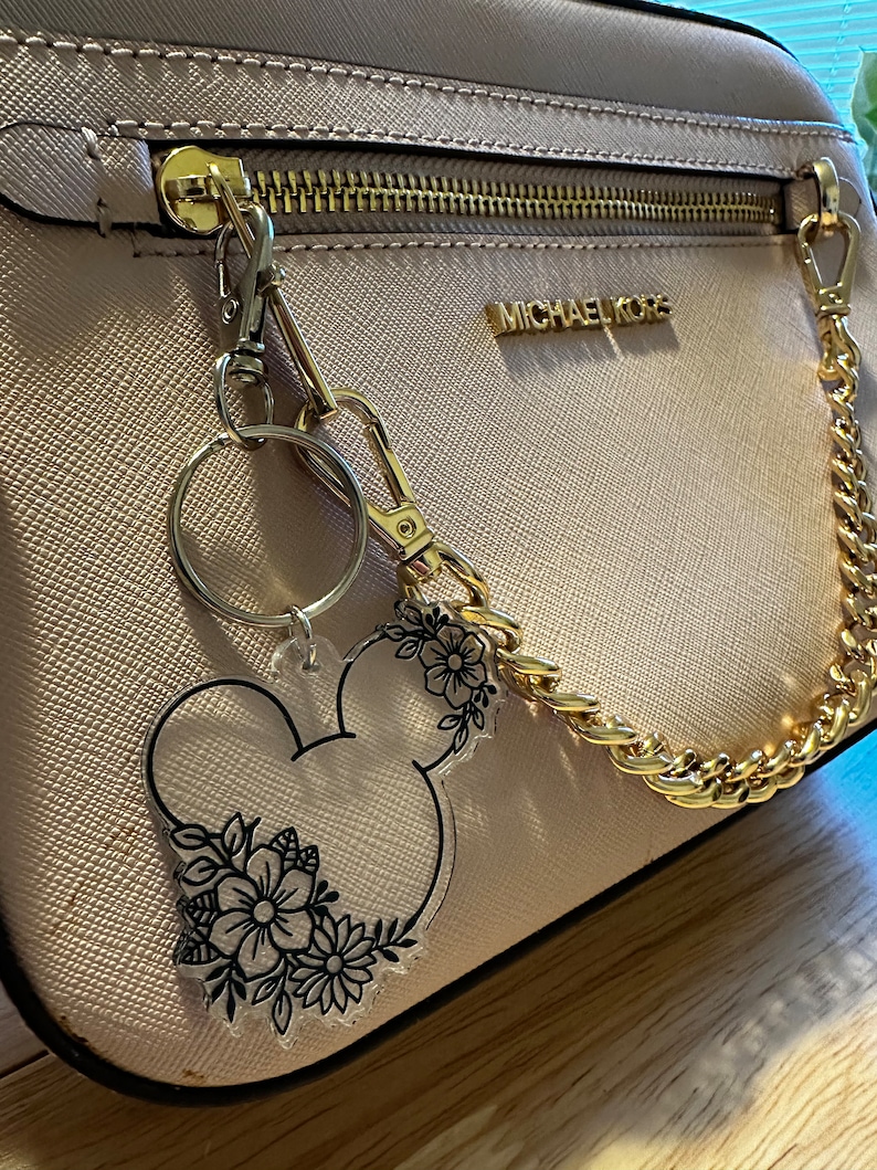 K&ouml;nnte beinhalten: Eine hellrosa Michael Kors Handtasche mit einer goldenen Kettenriemen und einem durchsichtigen Schl&uuml;sselanh&auml;nger mit einer schwarzen Umrandung von Mickey Mouse Kopf mit floralen Details.