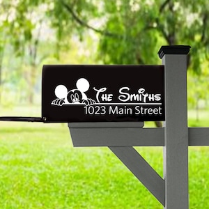 May include: Black mailbox with white text and a Mickey Mouse graphic. The text reads "The Smiths" and "1023 Main Street". The mailbox is mounted on a gray post with a green grassy background and a blurred tree line.