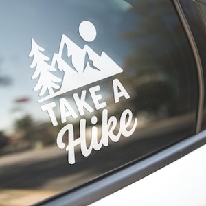 Puede incluir: Calcomanía blanca con las palabras "Take a Hike" en una fuente estilizada, acompañada de una cordillera, un sol y un diseño de pino. La calcomanía está adherida a la ventana de un coche.