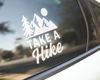 May include: White decal with the words "Take a Hike" in a stylized font, accompanied by a mountain range, a sun, and a pine tree design. The decal is affixed to a car window.