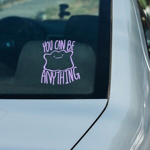 May include: A light purple vinyl decal applied to a car window. The design features a simple, blob-like character with a small face, positioned above and below the text "YOU CAN BE ANYTHING" written in a casual, hand-lettered style font.